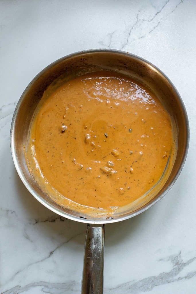A silver saucepan on a marble countertop containing thick, creamy orange sauce with small chunks, herbs, and spices.