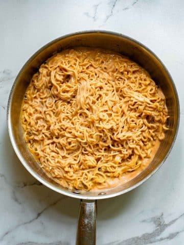 A skillet filled with creamy ramen noodles, showing a rich, golden-brown sauce coating the strands. The skillet is placed on a light marble countertop.