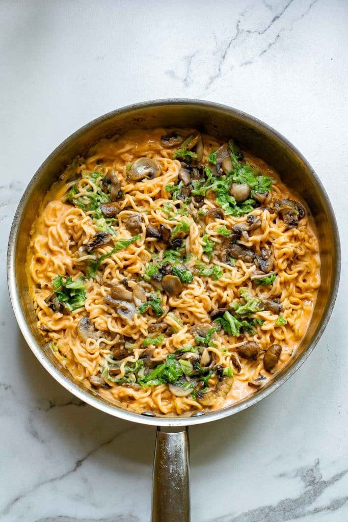 A pan filled with creamy ramen noodles topped with sliced mushrooms and garnished with fresh green herbs on a marble countertop.