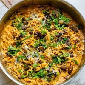 A skillet filled with creamy noodles topped with sliced mushrooms, chopped green onions, and grated cheese, garnished with fresh parsley. The dish has a rich, savory appearance.