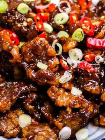 A plate of crispy Mongolian beef garnished with sliced green onions and red chili peppers. The beef is glazed in a dark, glossy sauce, and is served on a patterned ceramic dish.