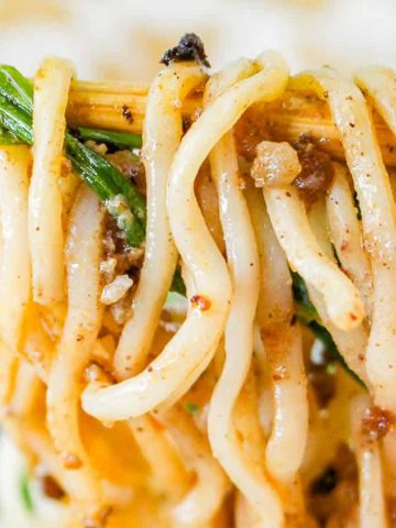 Close-up of chopsticks holding noodles coated in a savory sauce, garnished with chopped greens and bits of herbs. The background shows a blurred bowl of noodles.
