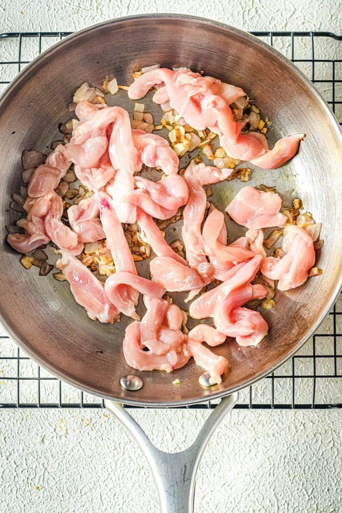 Sliced raw chicken and chopped onions in a frying pan on a wire rack, ready to be cooked. The shiny metal pan contrasts with the textured white surface underneath.