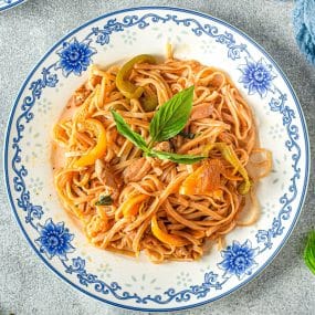 A decorative plate filled with noodles, slices of bell pepper, and meat, garnished with fresh basil. The blue and white floral pattern on the plate contrasts with the vibrant colors of the food. The plate is set on a gray surface.