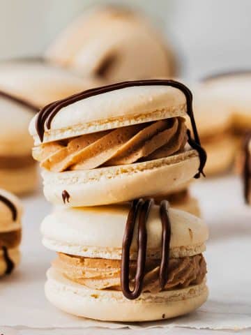 A stack of French macarons filled with creamy chocolate ganache and drizzled with dark chocolate on parchment paper. Other macarons are scattered in the background, creating a delicious, elegant arrangement.