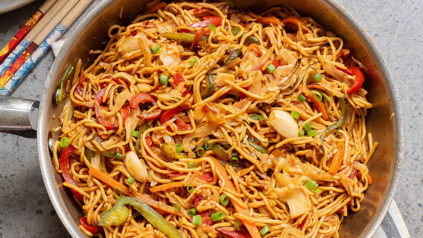 A pan filled with stir-fried hakka noodles mixed with sliced vegetables such as bell peppers, onions, green onions, and cabbage, garnished with chopped green onions. Chopsticks are placed beside the pan.