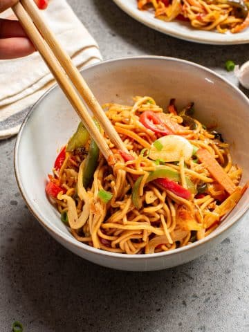 A bowl of stir-fried noodles with colorful vegetables like bell peppers and scallions. A hand uses chopsticks to pick up the noodles. The dish is served in a white bowl, set on a gray tabletop. Another bowl is partially visible in the background.
