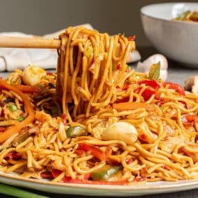 A close-up of a plate of stir-fried noodles with vegetables, including red bell peppers, carrots, and green onions. Mushrooms are also visible, and chopsticks are lifting some noodles from the dish.