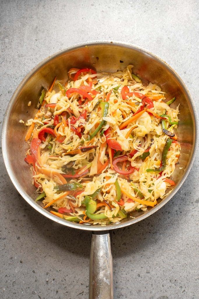 A stainless steel pan filled with a stir-fry of shredded cabbage, sliced red and green bell peppers, and thin carrot strips, all sautéed and lightly browned. The pan sits on a speckled gray countertop.