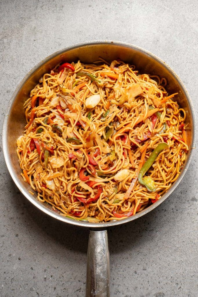 A pan of stir-fried noodles with sliced red and green bell peppers, onions, and other vegetables, mixed with a savory sauce on a gray surface.