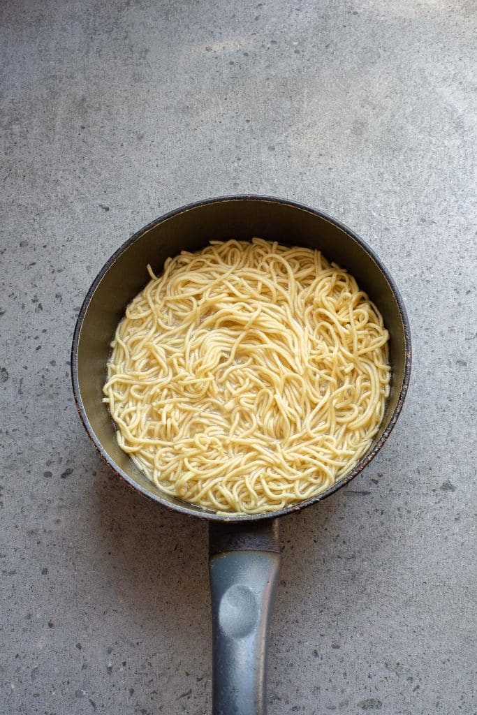 A black saucepan filled with cooked noodles sits on a gray speckled countertop.