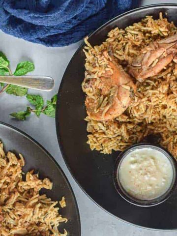 Three black plates filled with chicken and rice biryani are arranged on a table. Each plate is accompanied by a small bowl of creamy white sauce. Fresh mint leaves are placed beside the plates, and a dark blue cloth napkin is partially visible.