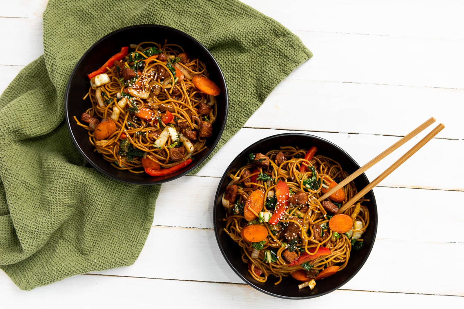 Two black bowls of stir-fried noodles with vegetables and meat, garnished with sesame seeds, sit on a white wooden table. A pair of chopsticks rests on one bowl. A green fabric napkin is partially visible under the bowls.