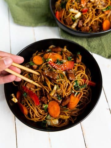 A bowl of stir-fried noodles with mixed vegetables and pieces of meat, held by a person using chopsticks. The vibrant dish includes carrots, red bell peppers, onions, and green leafy vegetables, garnished with sesame seeds. Another bowl is visible in the background.
