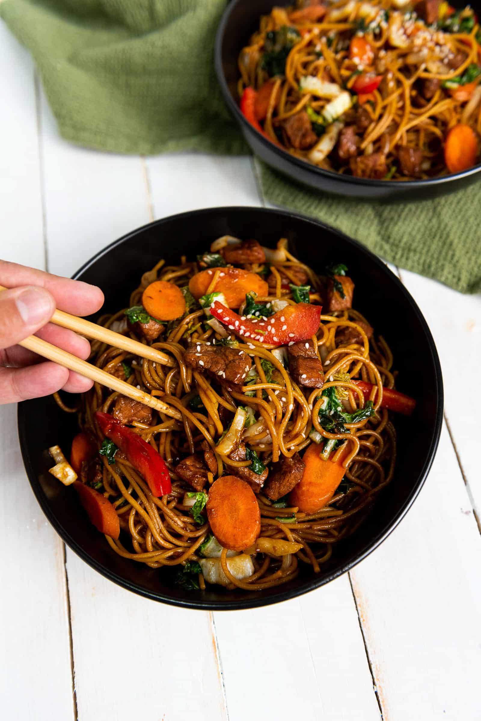 A bowl of stir-fried noodles with mixed vegetables and pieces of meat, held by a person using chopsticks. The vibrant dish includes carrots, red bell peppers, onions, and green leafy vegetables, garnished with sesame seeds. Another bowl is visible in the background.