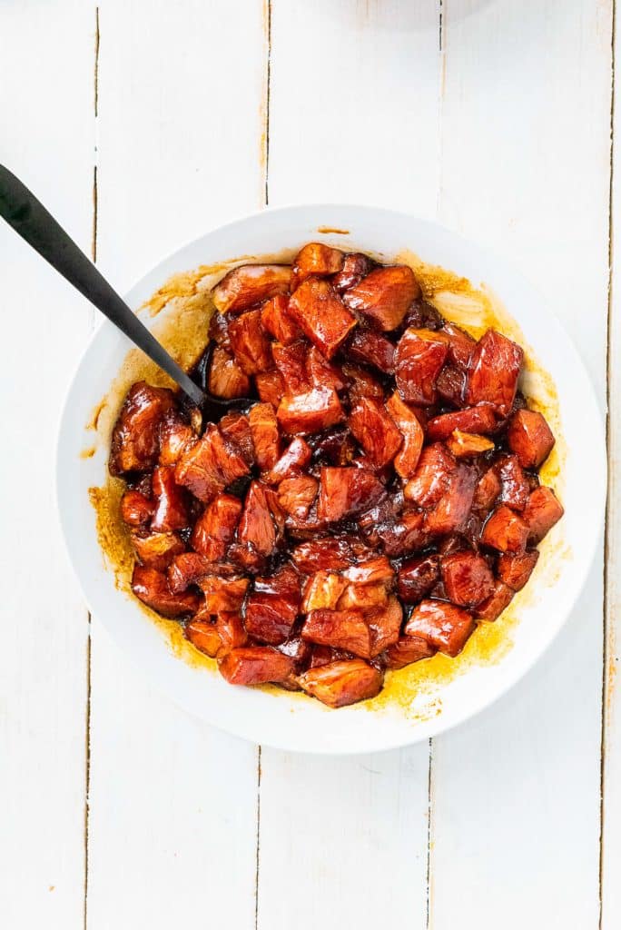 A white bowl filled with browned, cubed pieces of pork marinated in a savory sauce, with a black spoon resting on the left side of the bowl. The bowl is placed on a white wooden surface.