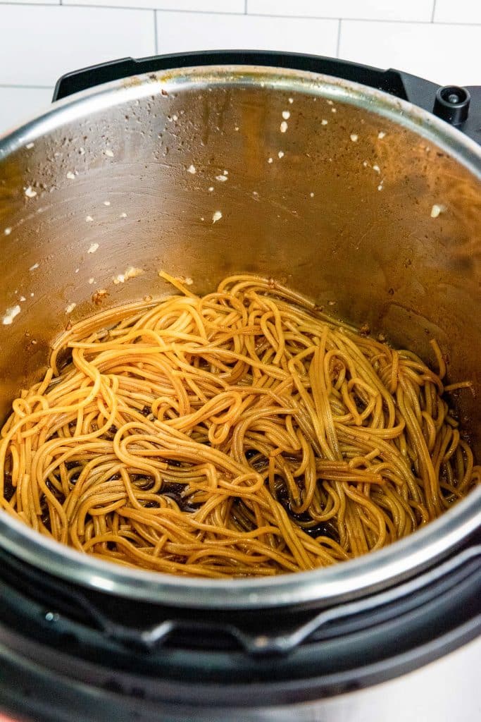 An inner pot of an Instant Pot filled with cooked spaghetti coated in a light brown sauce. The pot is stainless steel, and the background shows white subway tiles. Some sauce residue is visible on the sides of the pot.
