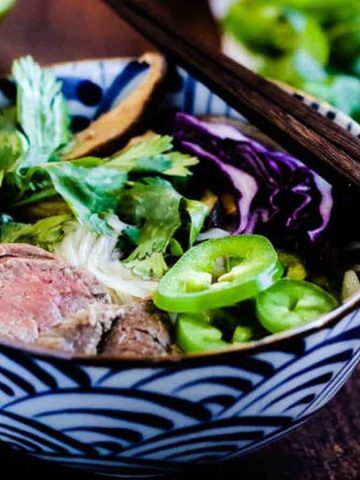 A colorful bowl of ramen is topped with sliced beef, lime wedges, fresh greens, jalapeño slices, purple cabbage, and mushrooms. Chopsticks rest on the bowl's rim, and a halved lime is visible in the background.