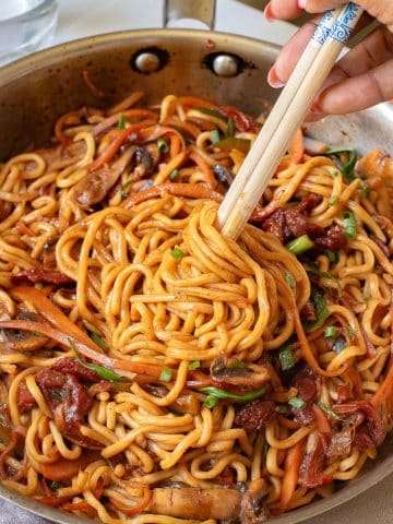 A person uses chopsticks to pick up a serving of stir-fried noodles from a pan. The dish includes carrots, mushrooms, and green onions, all cooked in a savory sauce.