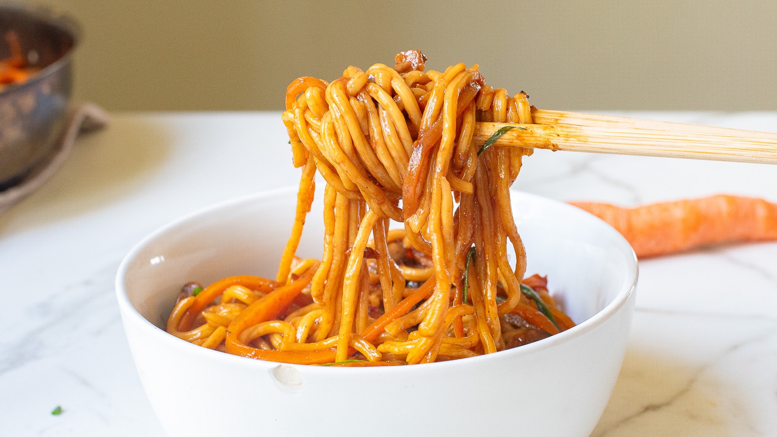 A bowl of saucy noodles is being lifted with chopsticks over a marble countertop. A carrot is partially visible in the background.