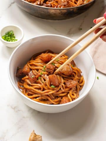A bowl of Asian-style noodles with chunks of meat, garnished with chopped green onions. A hand holds chopsticks, picking up noodles. A small dish with green onions and a piece of ginger are nearby on a white surface.