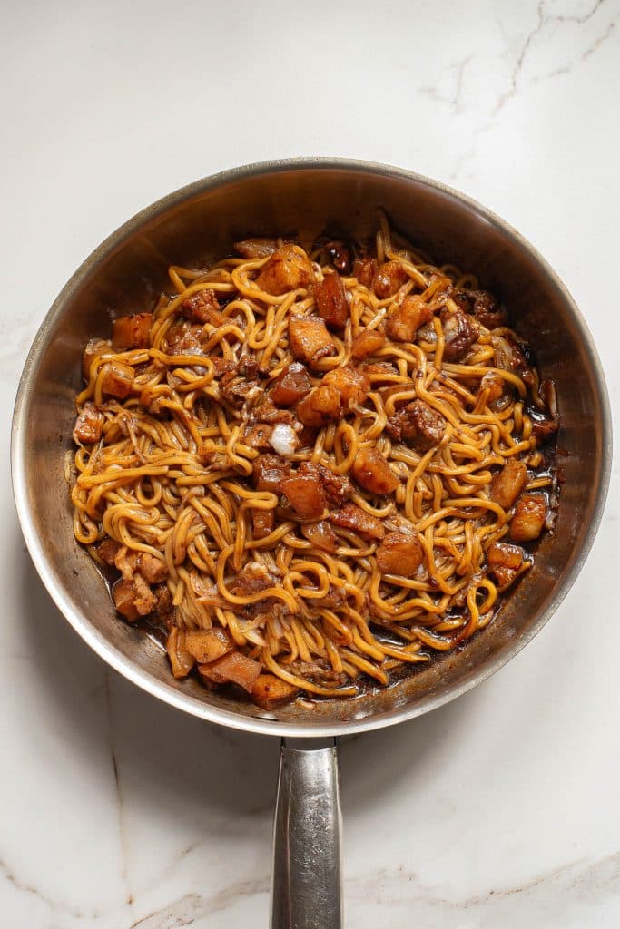 A stainless steel pan filled with stir-fried noodles and diced chicken, coated in a glossy sauce, placed on a white marble surface.