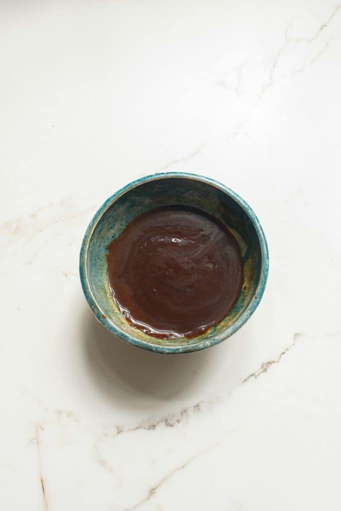 A small blue-green ceramic bowl containing a smooth, dark brown sauce sits on a marbled white surface. The bowl's interior texture is slightly visible under the sauce.