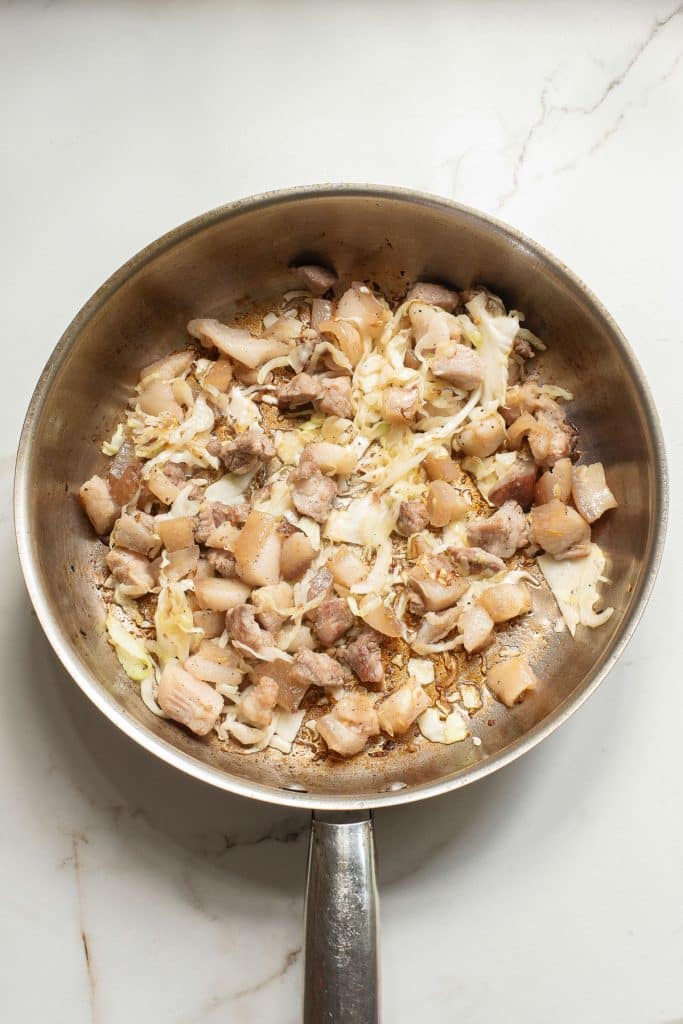 A frying pan on a marble countertop contains diced bacon and shredded cabbage, slightly browned and cooked together. The ingredients are spread evenly across the pan.