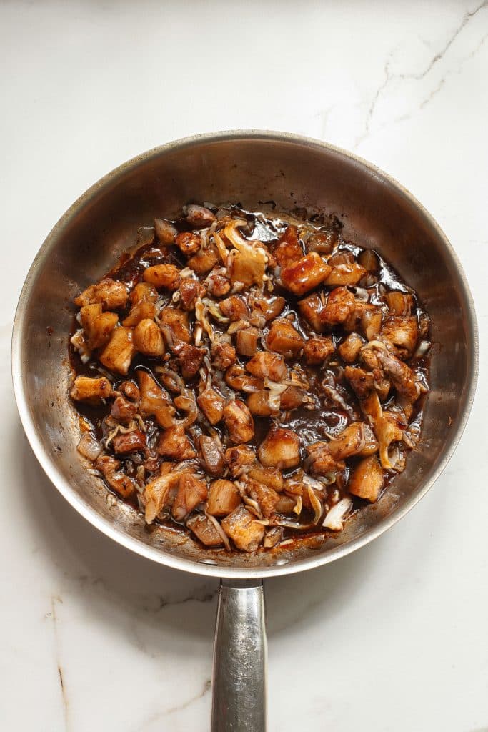 Frying pan with cooked cubed chicken and onions in a brown sauce on a light marble surface.