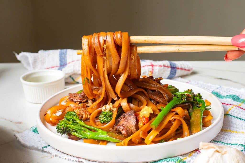 A plate of stir-fried noodles with vegetables like broccoli and carrots. A hand is holding chopsticks, lifting some noodles. The background features a colorful cloth and a small white sauce dish.
