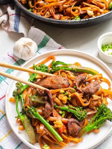 A plate of stir-fried noodles with sliced beef, broccolini, and vegetables, garnished with chopped scallions. Chopsticks rest on the plate. A pan with more of the dish, a garlic bulb, and a small bowl of scallions are in the background.