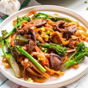 A plate of pad see ew with stir-fried noodles, tender beef slices, broccolini, and mixed vegetables, all elegantly garnished with sauce. The dish rests on a white plate while a garlic bulb sits partially visible in the background.