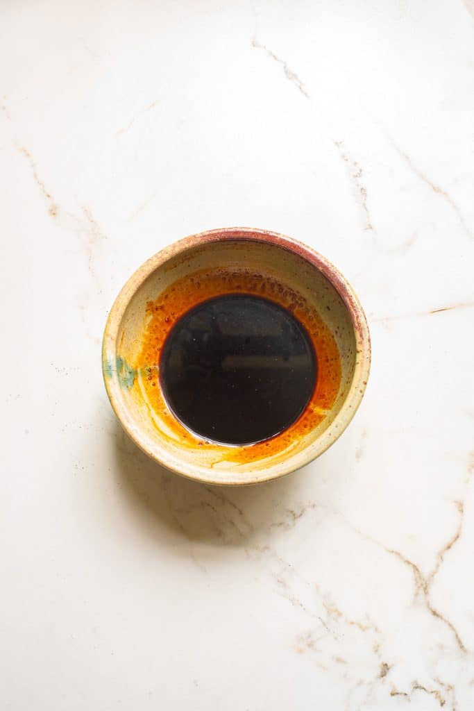 A ceramic bowl sits on a marble surface, filled with a dark soy sauce. The bowl has earthy tones and a smooth texture, contrasting with the glossy liquid inside.