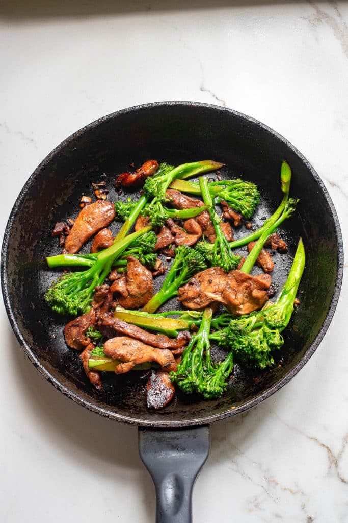 A black skillet contains stir-fried chicken pieces and vibrant green broccolini on a light-colored marble surface. The dish is seasoned, giving the chicken a glazed appearance. Shadows cast on the left side add depth to the image.