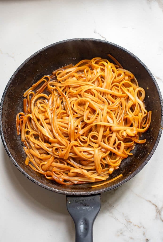 A frying pan filled with cooked linguine pasta coated in a light brown sauce, positioned on a white marble surface.