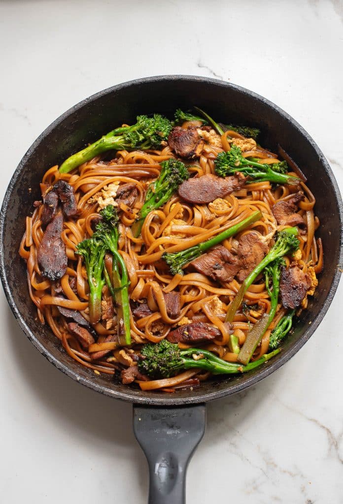 A pan filled with stir-fried noodles, sliced beef, and broccolini. The dish is seasoned and cooked with a sauce, giving it a rich, glossy appearance. It rests on a marble countertop.
