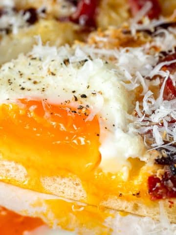 A pizza slice topped with a runny fried egg, shredded cheese, pepper, and small pieces of meat on a golden crust. The yolk is visibly glossy and slightly cracked, oozing over the slice.