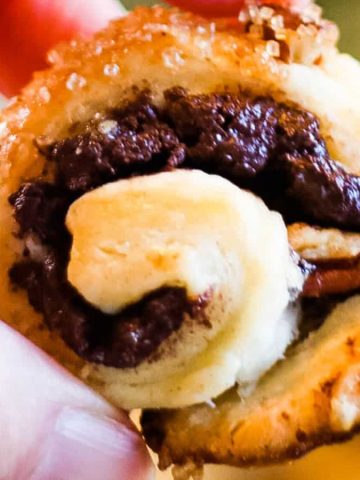 Close-up of a hand holding a freshly baked pastry roll filled with melted chocolate and nuts. The pastry is golden brown with a flaky texture, and the filling is dark and glossy, contrasting with the light, crispy dough.