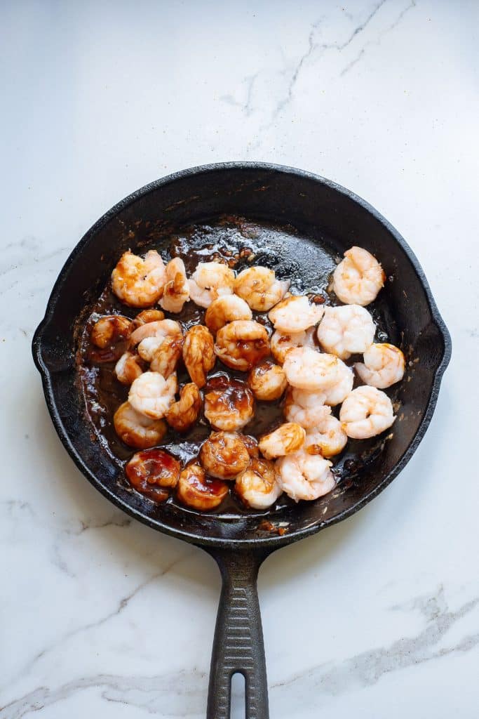 A black skillet on a white marble surface contains shrimp being sautéed. Some shrimp are pink and cooked, while others are partially cooked with a raw, translucent appearance. The shrimp are coated in a glossy brown sauce.