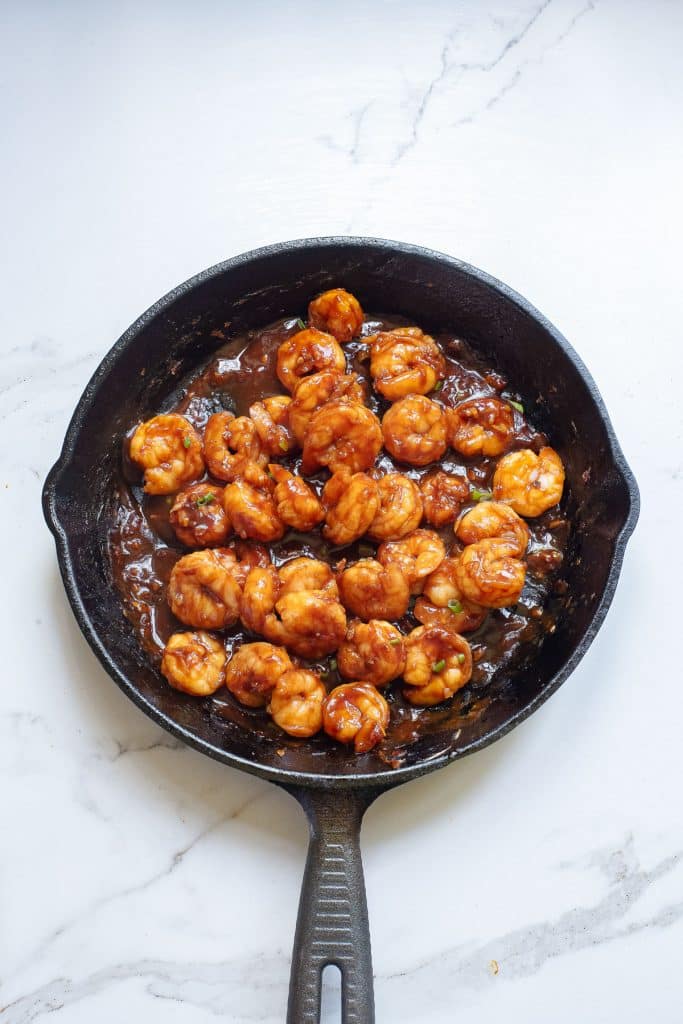 A cast iron skillet filled with cooked shrimp coated in a rich, brown sauce, placed on a white marble countertop. The shrimp are evenly distributed and glistening with the sauce, showcasing a savory, appetizing dish.