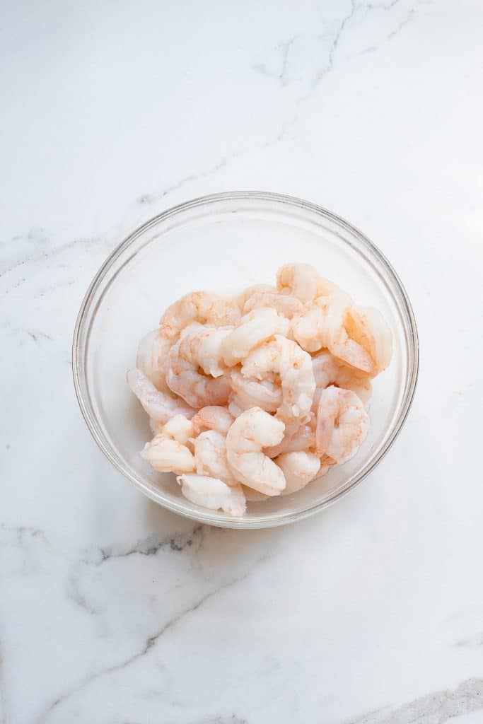 A clear glass bowl filled with raw, peeled shrimp sits on a white marble countertop. The shrimp are light pink and white, indicating they are uncooked. The background is clean and minimalist, focusing attention on the bowl of shrimp.