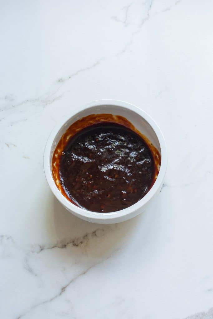 A white bowl filled with a thick, dark brown sauce sits on a white marble surface. The sauce appears smooth with a glossy texture, indicative of rich flavor.