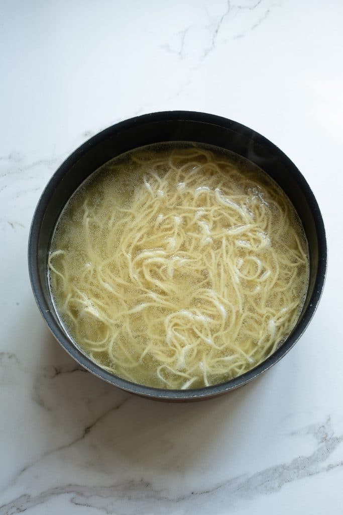 A black bowl filled with noodles in a clear broth sits on a white marble surface. The noodles are tangled and submerged in the liquid, which appears to be a simple, light-colored soup base.