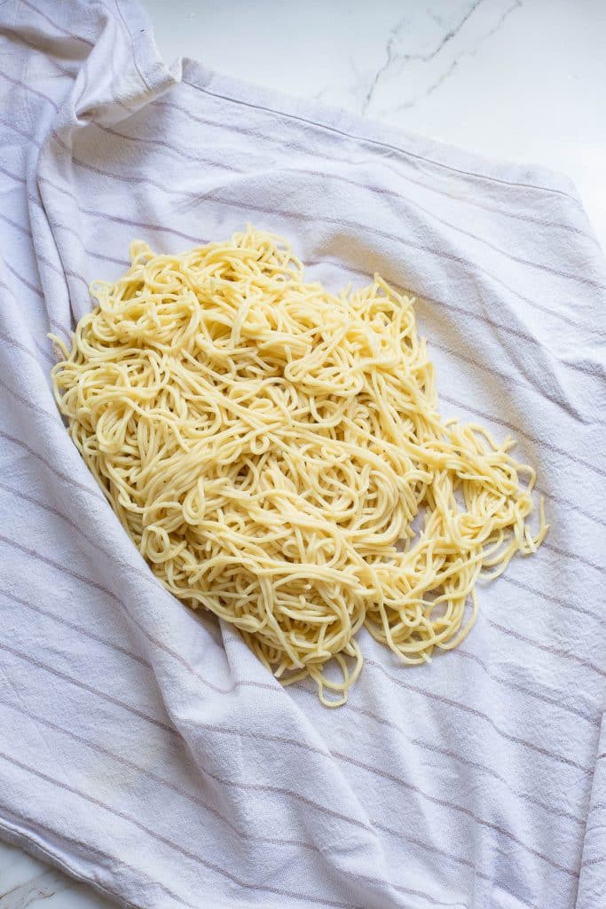 A large portion of cooked yellow noodles is spread out on a white cloth with faint gray stripes, laid on a white marble countertop. The noodles appear to be freshly made, with a soft texture.