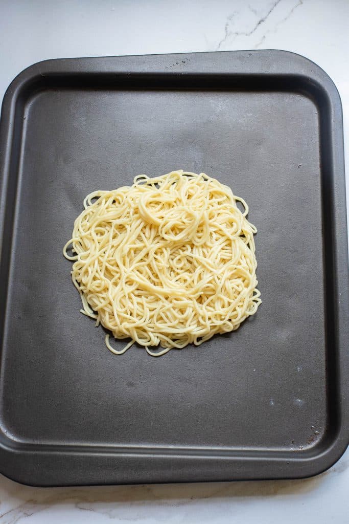 A pile of uncooked spaghetti noodles is spread out on a dark metal baking sheet. The edges of the baking sheet are slightly raised, and the surface appears clean and smooth. The background is a light marble countertop.
