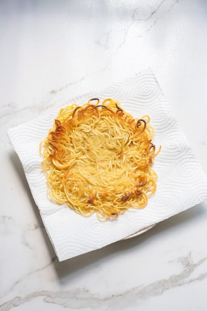 A golden brown, crispy round ramen patty placed on a white paper towel, which is on a white marble surface. The ramen patty has slightly charred edges, indicating it has been fried.