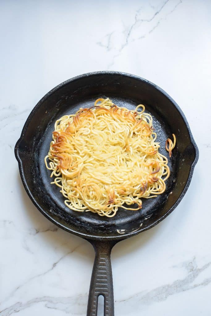 A cast iron skillet containing a lightly browned nest of cooked noodles sits on a white marble countertop. The noodles are golden and crisp around the edges, forming a circular pattern.
