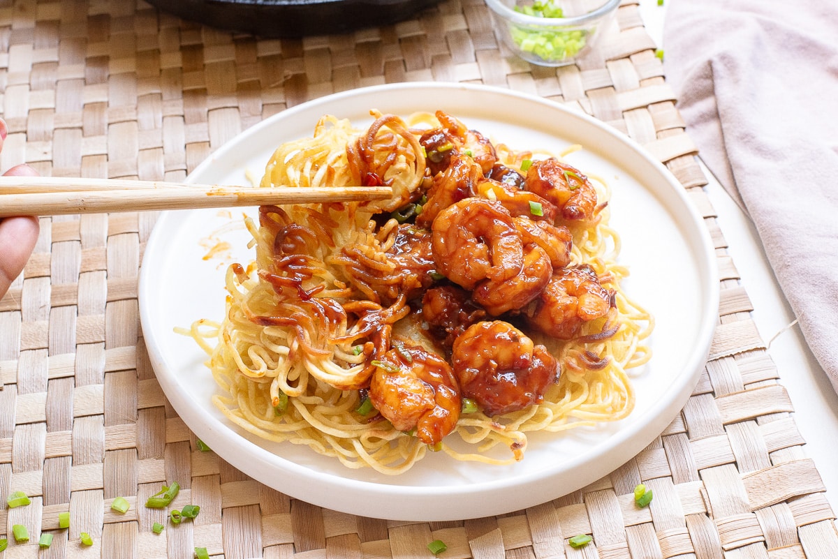 A white plate holding a serving of crispy noodles topped with saucy shrimp, garnished with green onions. A pair of chopsticks is lifting a portion of the noodles and shrimp. The plate is on a woven mat, and a small dish with green onions is visible in the background.