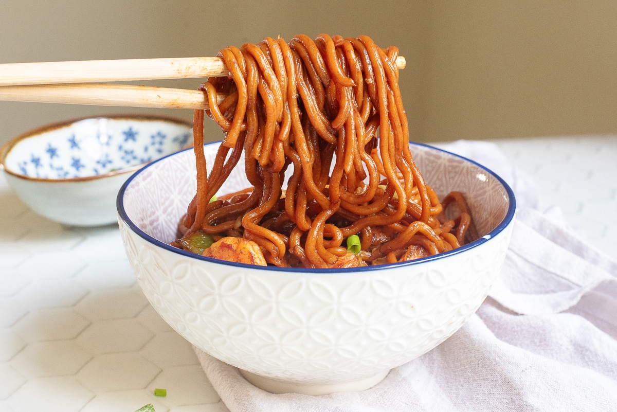 A bowl of spicy noodles with chopsticks lifting a portion. The noodles are coated in a rich red sauce, garnished with green onions. The bowl has a textured white design, and a small blue and white patterned bowl is in the background.