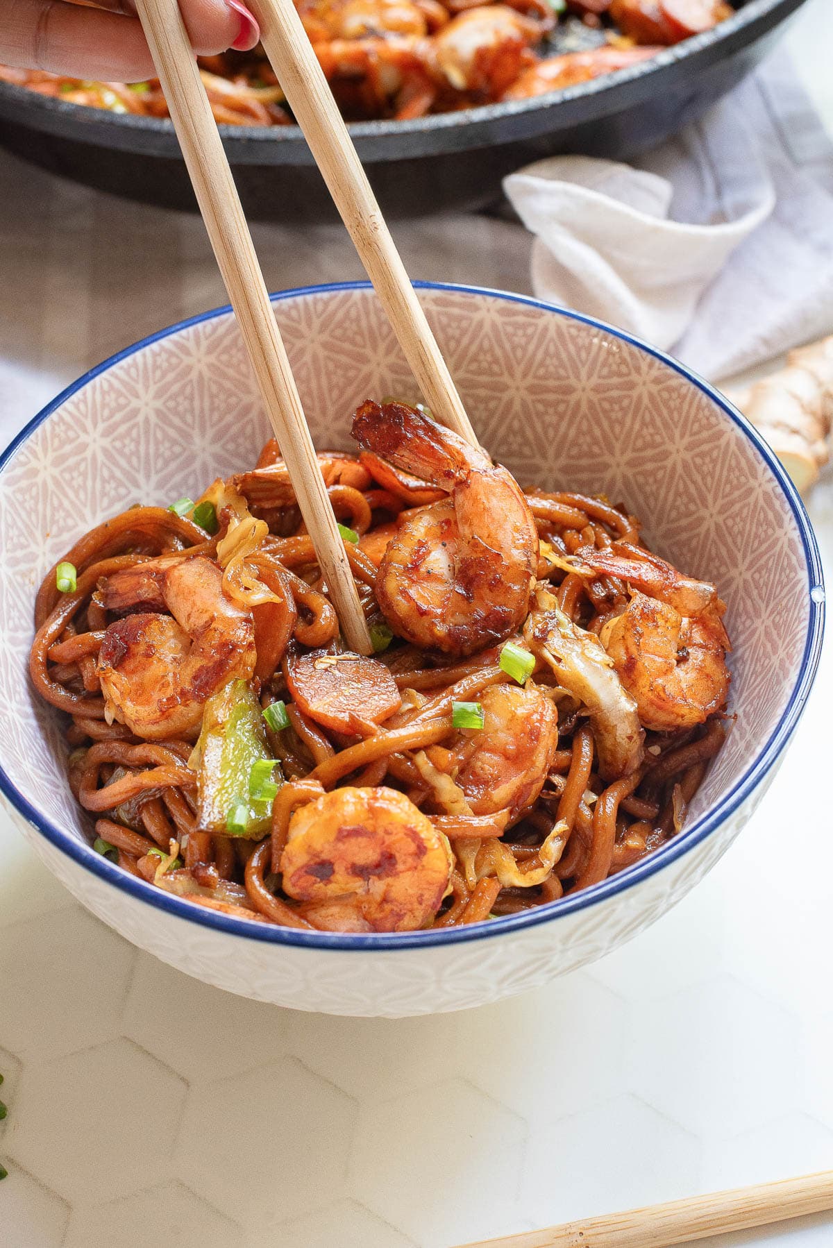 A bowl of noodles with shrimp and vegetables is held by hand using chopsticks. The dish features a savory sauce and is garnished with green onions. A skillet with more food is visible in the background, resting on a white surface.